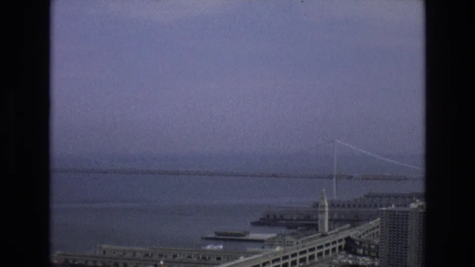 1973 San Francisco home-movie frame showing a bridge crossing the bay, waterfront buildings, roadways, and pale haze over the water.