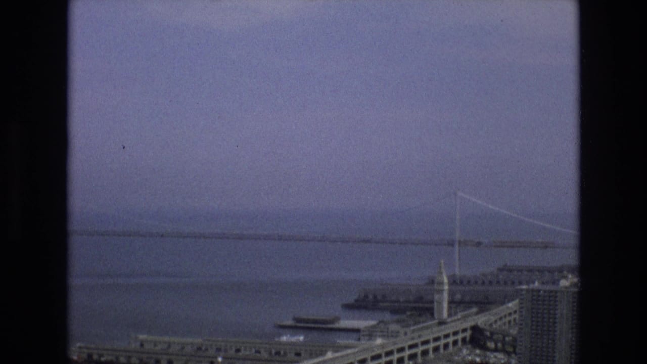 1973 8mm view from San Francisco showing a hazy bay, a distant suspension bridge, piers, waterfront buildings, and a towered structure under an overcast sky.