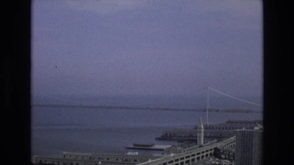 1973 8mm view from San Francisco showing a hazy bay, a distant suspension bridge, piers, waterfront buildings, and a towered structure under an overcast sky.