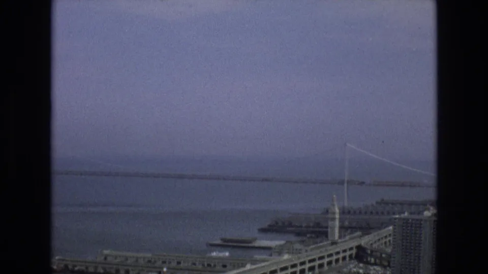 Hazy 1973 San Francisco bayfront frame with a distant suspension bridge, industrial-looking shoreline structures, piers, and muted blue-gray sky.