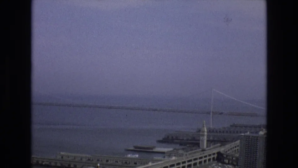 1973 8mm San Francisco waterfront view with the distant bridge partly softened by haze above bay water and low buildings near the shoreline.