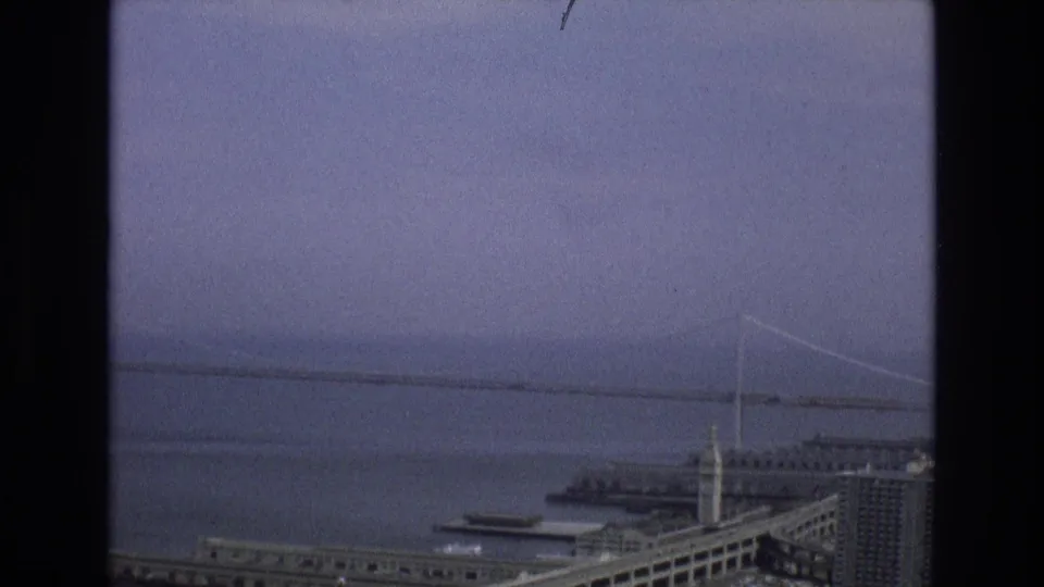 Final 1973 8mm frame holding on the San Francisco bayfront, with a distant suspension bridge, broad water surface, foreground piers, and dark frame borders.