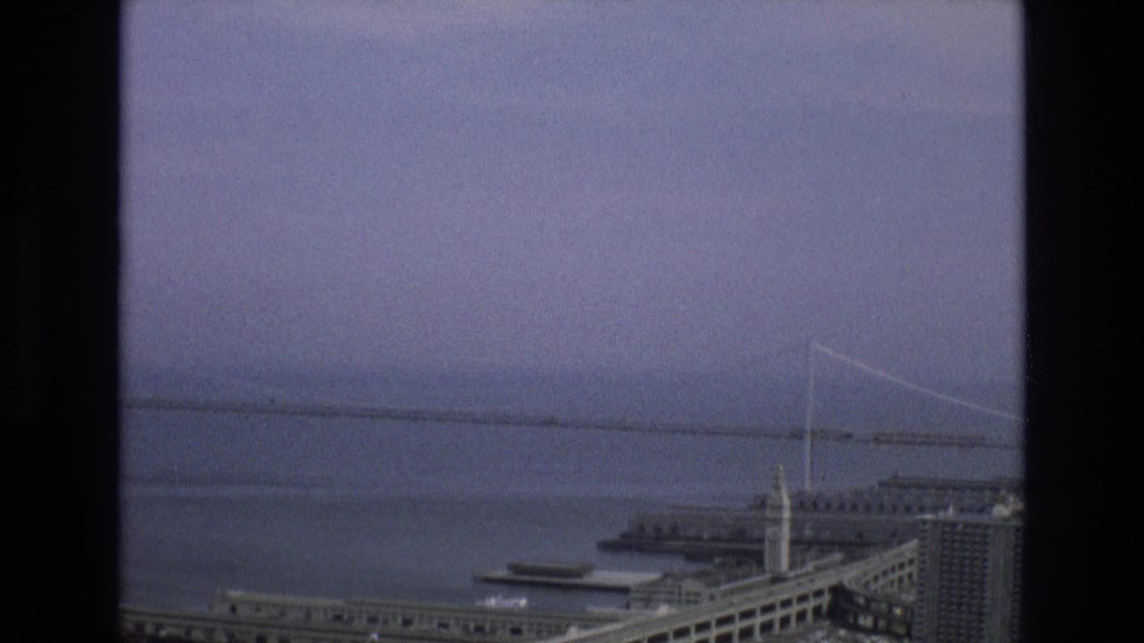 1973 San Francisco waterfront seen from an elevated interior perspective, with a long suspension bridge across the bay, a clock tower building, piers, and a curving roadway in the foreground.