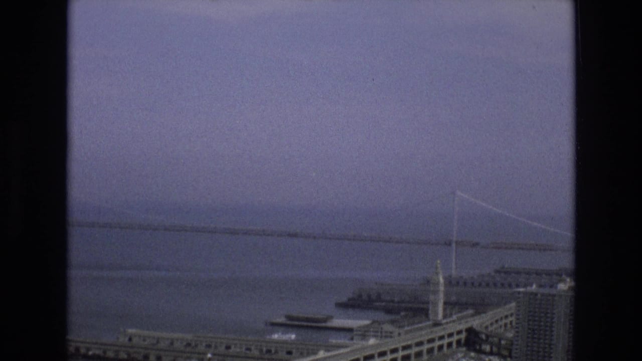 Elevated 1973 view over San Francisco's waterfront, showing bay water, a distant suspension bridge, a clock tower, piers, and urban buildings beneath a muted sky.