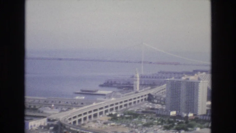 Brightly exposed 8mm frame of the 1973 San Francisco waterfront, with bridge cables crossing the bay, a clock tower, piers, buildings, and a sweeping elevated roadway.
