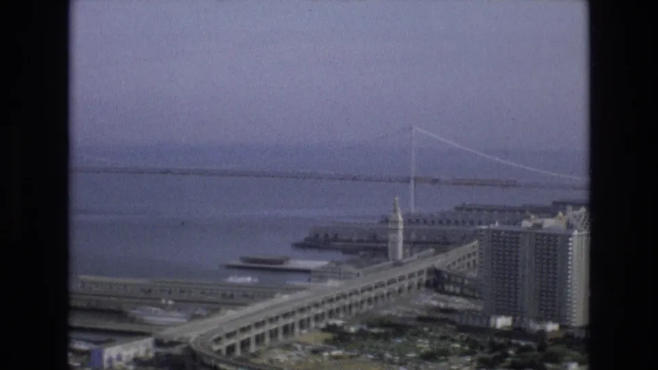 1973 San Francisco bayfront view from above, showing a curving elevated roadway, clock tower building, waterfront structures, bay water, and a distant suspension bridge in haze.