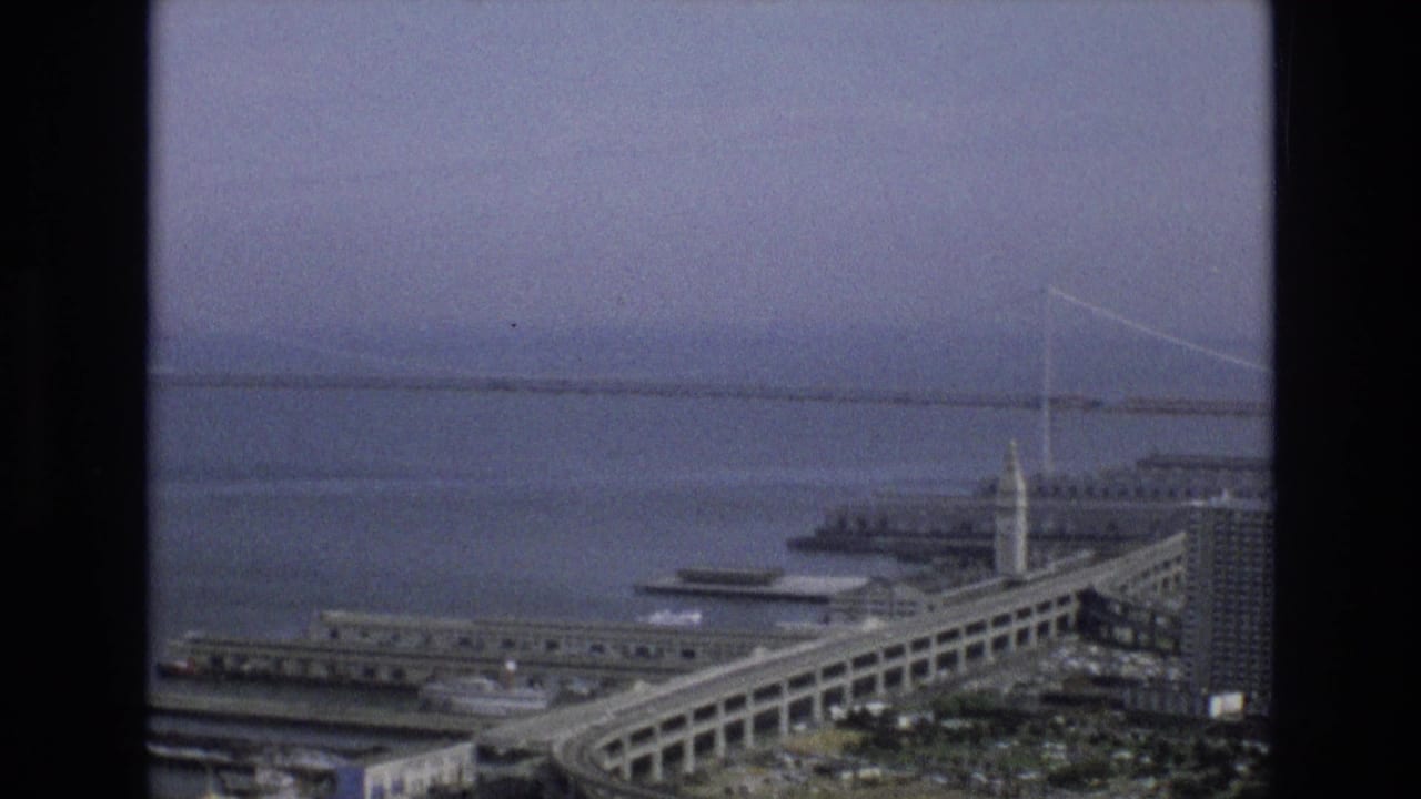1973 San Francisco aerial view showing the Bay Bridge across the bay, waterfront piers, a ferry or boat, and the Ferry Building tower near the Embarcadero.
