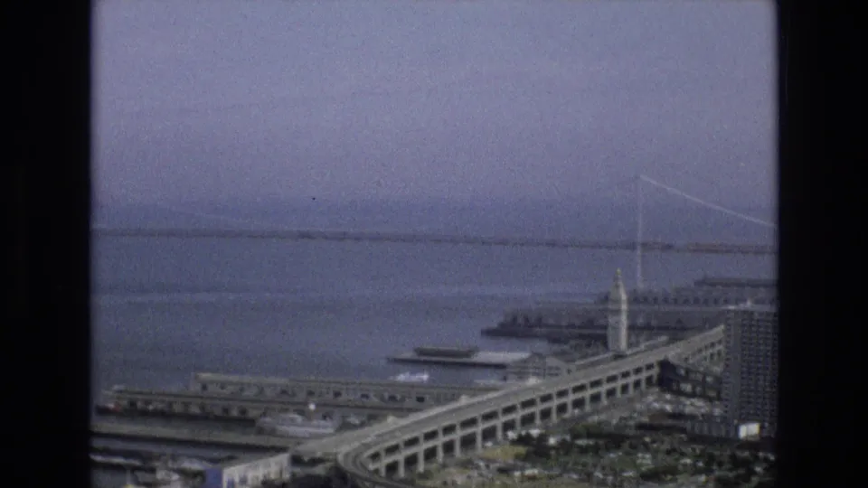 1973 San Francisco aerial view showing the Bay Bridge across the bay, waterfront piers, a ferry or boat, and the Ferry Building tower near the Embarcadero.