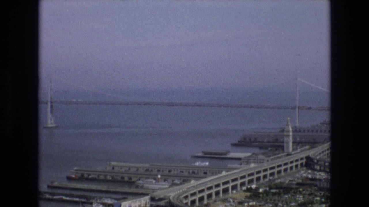 Aerial 8mm frame of San Francisco's waterfront with the Bay Bridge in the background, piers along the Embarcadero, bay water, and the Ferry Building tower visible.