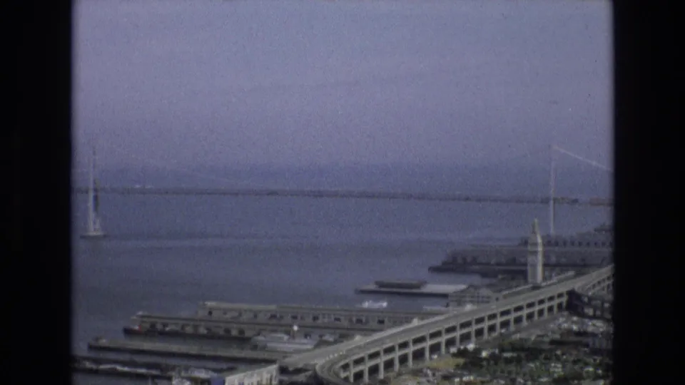 Aerial 8mm frame of San Francisco's waterfront with the Bay Bridge in the background, piers along the Embarcadero, bay water, and the Ferry Building tower visible.