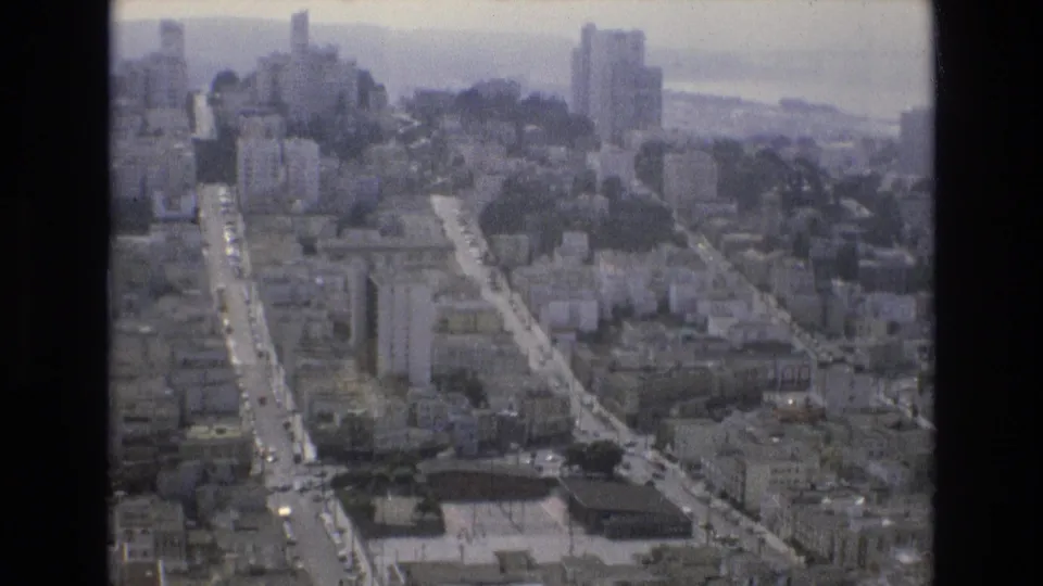 1973 San Francisco aerial frame looking across dense city blocks, multi-story buildings, traffic-lined streets, and distant bay water through a soft 8mm scan.