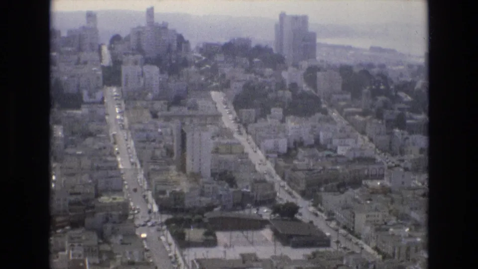 Aerial home-movie view of San Francisco in 1973, showing neighborhood blocks, traffic on straight streets, taller buildings, hills, and bay water in the distance.