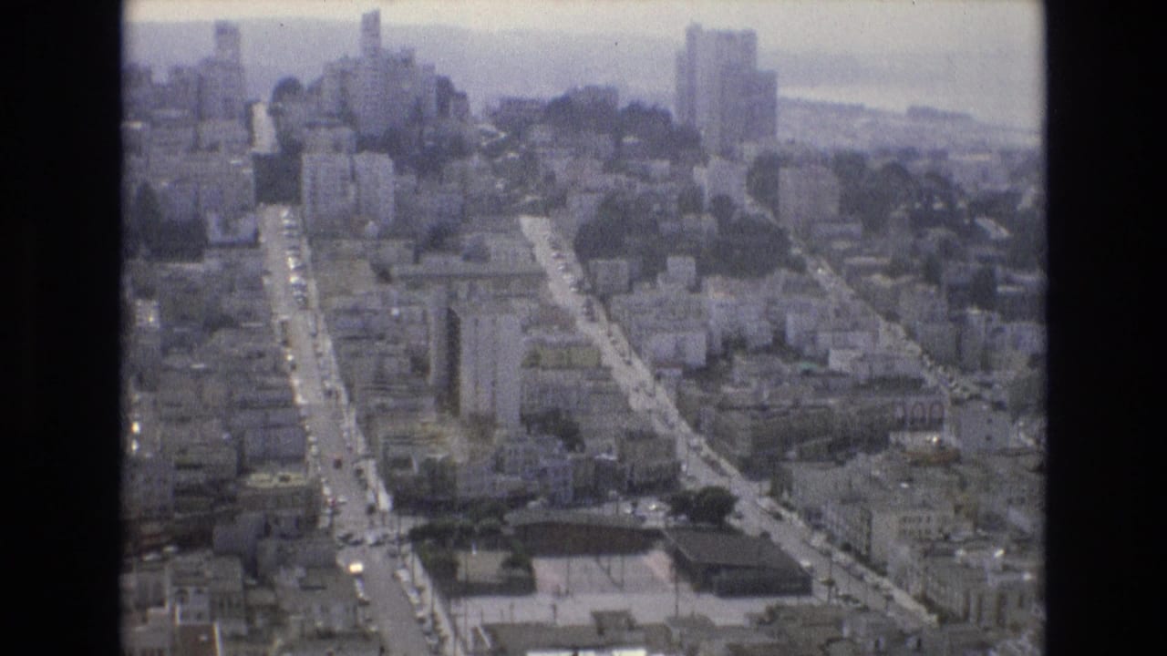 1973 San Francisco aerial home-movie frame looking over dense hillside streets, mid-rise buildings, taller towers, and hazy bay water in the distance.
