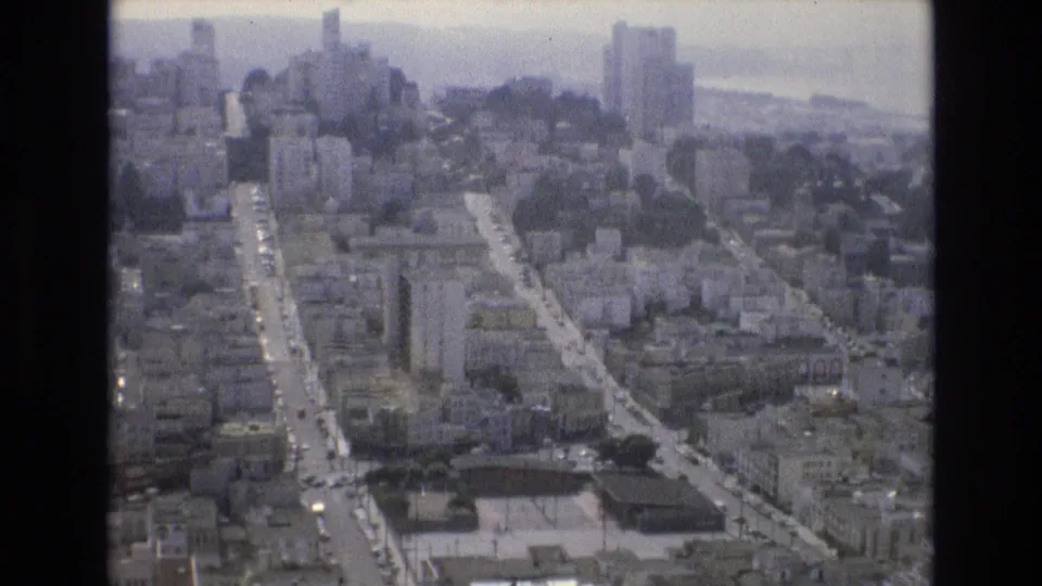 1973 San Francisco aerial home-movie frame looking over dense hillside streets, mid-rise buildings, taller towers, and hazy bay water in the distance.