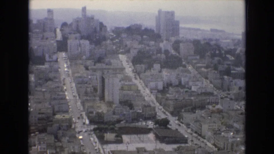 1973 San Francisco home-movie frame looking down a prominent hill street through tightly packed buildings, with taller structures and bay haze beyond.