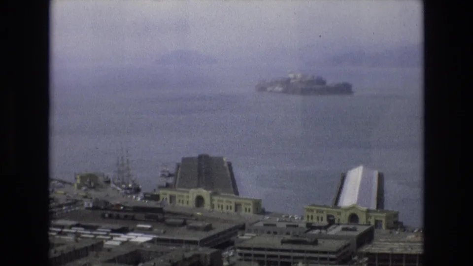 1973 San Francisco bay view with Alcatraz Island visible through haze beyond waterfront buildings and rooflines in the foreground.