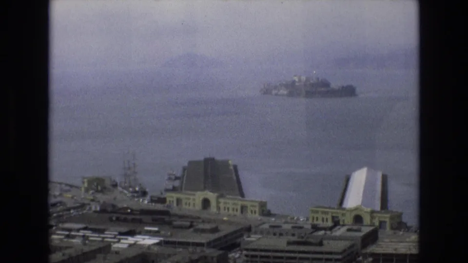 A hazy 1973 San Francisco waterfront view shows Alcatraz across the bay, with pier structures and ferry terminal architecture in the foreground.