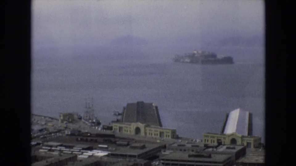 Soft, grainy 8mm footage frames San Francisco Bay with Alcatraz in the distance and Embarcadero waterfront buildings along the lower edge.
