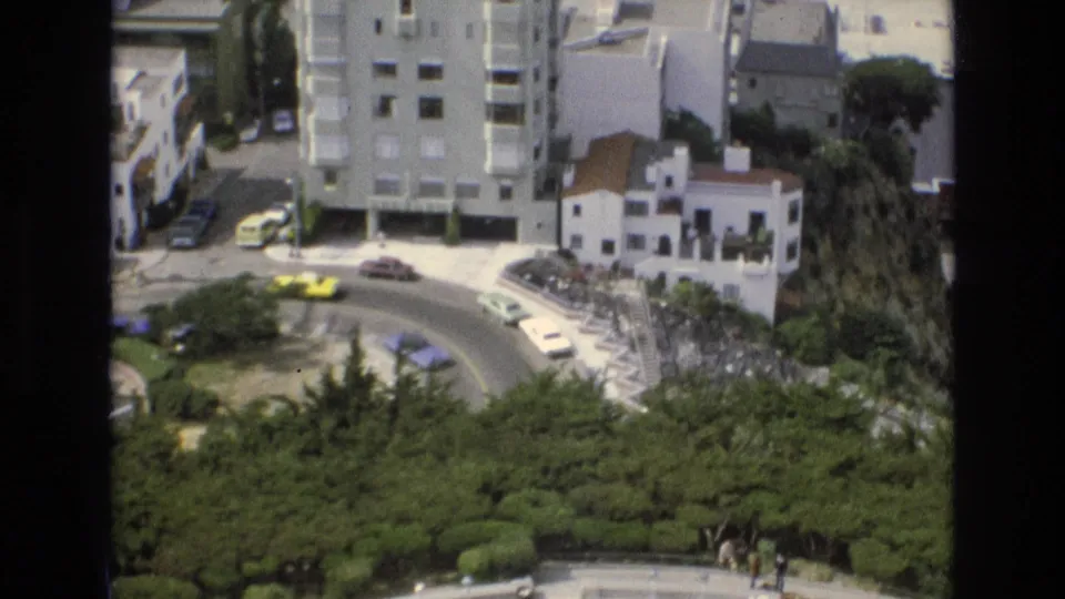 Elevated 1973 San Francisco street view showing a curved road, compact cars, white buildings, and landscaped greenery below the camera position.