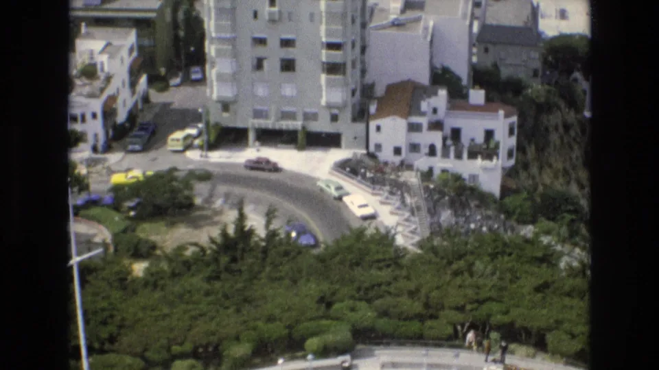 1973 San Francisco elevated street view with parked and moving cars around a bend, apartment-style buildings, sidewalks, and dense trees in the foreground.