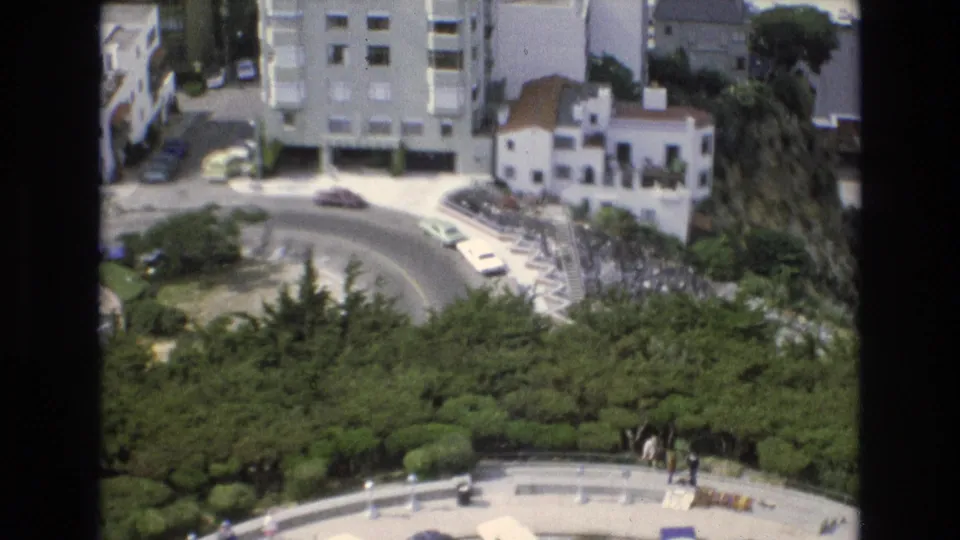 Elevated 1973 San Francisco view over a curved road, surrounding greenery, white buildings, parked cars, and small pedestrians near the lower walkway.