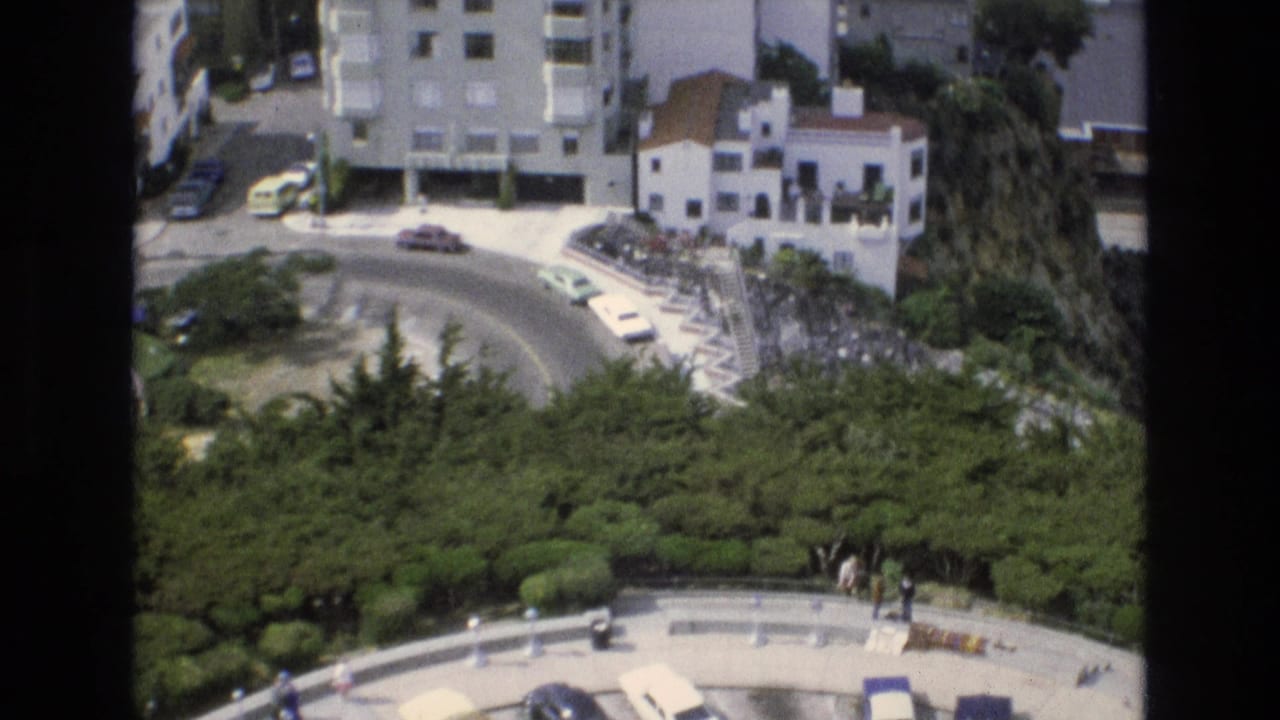 1973 San Francisco 8mm frame showing a curved hillside road beside parked cars, white residential buildings, dense greenery, and small pedestrians below.