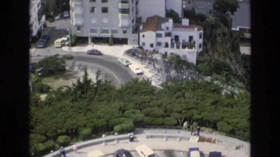 1973 San Francisco 8mm frame showing a curved hillside road beside parked cars, white residential buildings, dense greenery, and small pedestrians below.