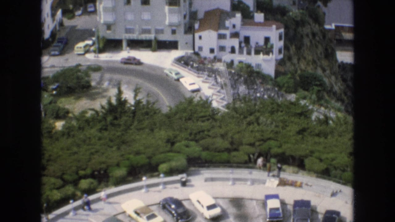 Curved roadway with cars, white hillside houses, trees, and people near the lower pavement.