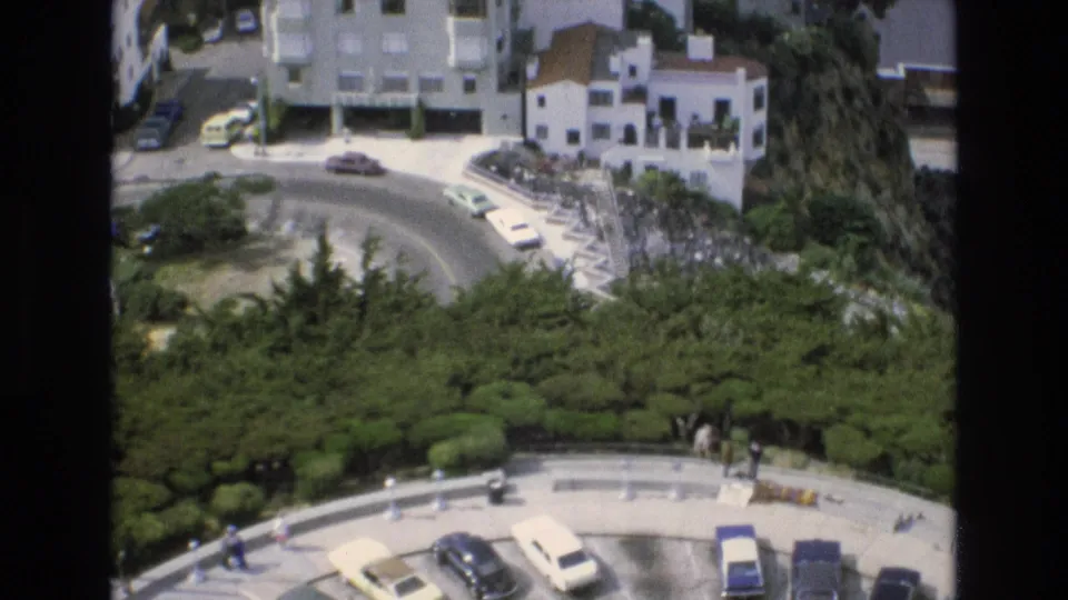 Elevated 1973 San Francisco view of a winding residential road, small parked cars, white hillside houses, greenery, and pedestrians along the lower edge.
