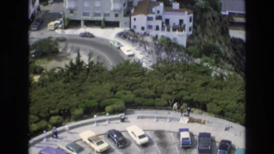1973 San Francisco home-movie frame with a lower parking lot, a curved road above it, period cars, hillside homes, greenery, and distant pedestrians.