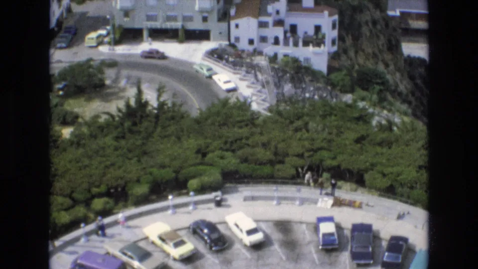 Final 1973 San Francisco frame showing parked cars in a lower lot, a curved road, hillside residential buildings, thick greenery, and small pedestrians in the distance.
