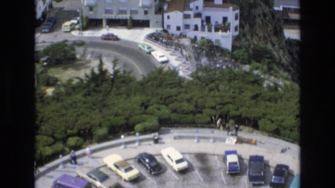 High-angle 1973 San Francisco home-movie view of a curved overlook road with parked cars, dense greenery, and white hillside buildings.