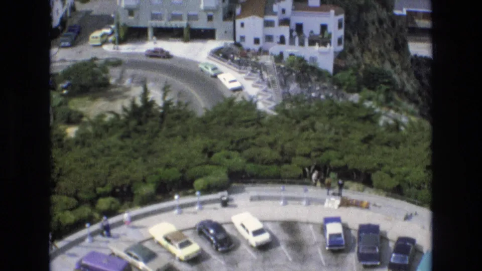 High-angle 1973 San Francisco home-movie view of a curved overlook road with parked cars, dense greenery, and white hillside buildings.