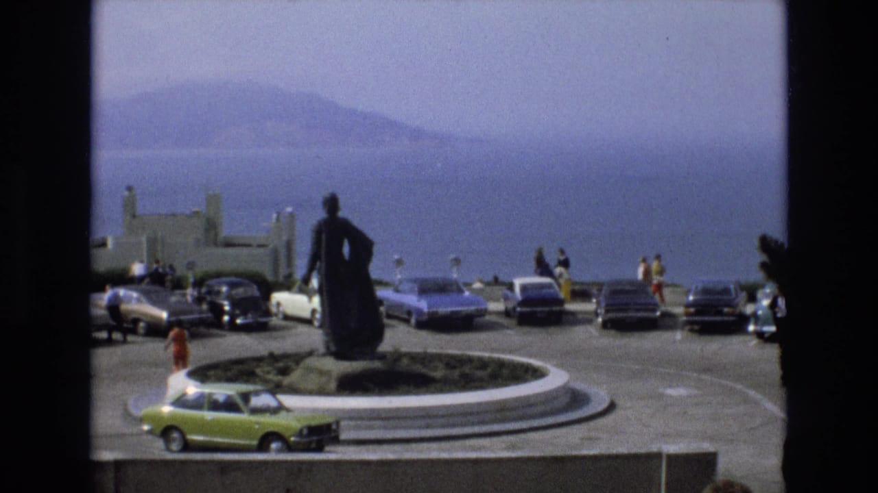 Statue on a circular traffic island with parked cars, visitors, water, and hazy hills beyond.