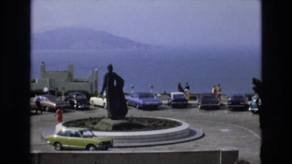 1973 San Francisco overlook scene with a statue in a circular traffic island, visitors near parked cars, and hazy water and hills in the background.