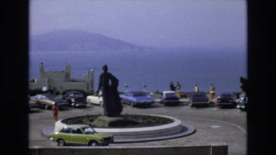 Wide 1973 San Francisco home-movie frame showing a statue at an overlook traffic circle, people standing near cars, and a hazy bay or ocean view.
