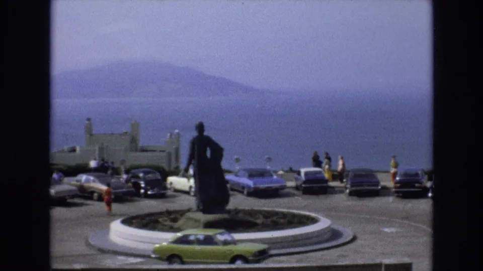 1973 San Francisco overlook footage frame with a bright green car passing the circular monument, visitors nearby, and blue water fading into distant hills.