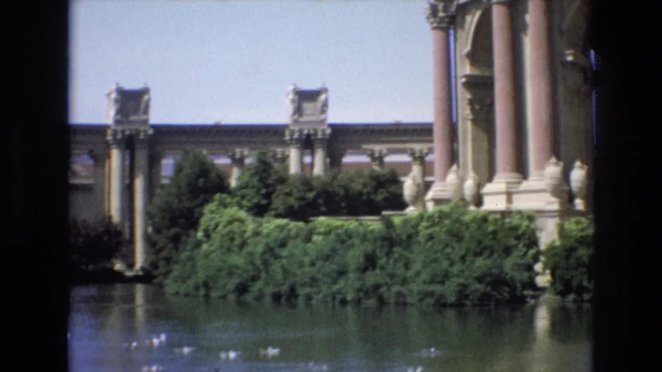 1973 8mm view of San Francisco's Palace of Fine Arts colonnade across a lagoon with ducks and dense greenery in the foreground.