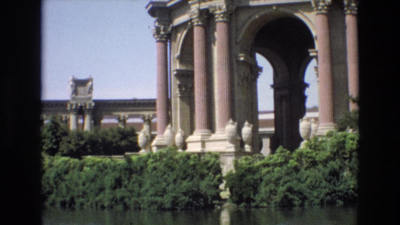 1973 8mm frame of the Palace of Fine Arts rotunda in San Francisco, showing pink columns, arched openings, hedges, and lagoon reflections.