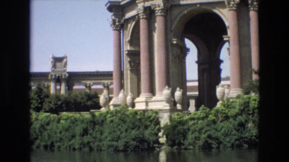 1973 8mm frame of the Palace of Fine Arts rotunda in San Francisco, showing pink columns, arched openings, hedges, and lagoon reflections.