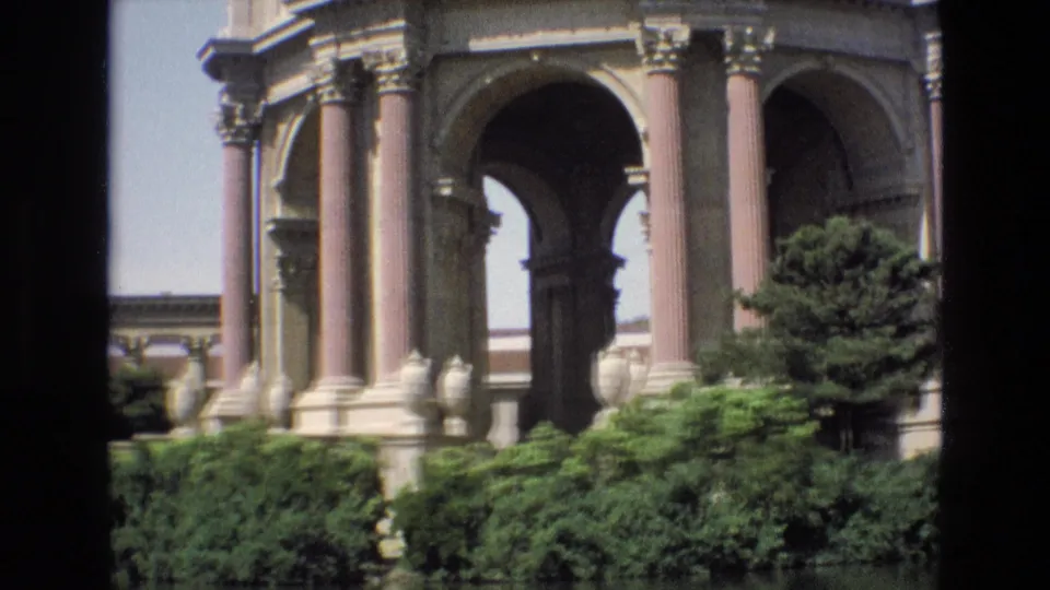 Closer 1973 home-movie view of the Palace of Fine Arts rotunda, with arched passages, columns, shrubs, a tree, and lagoon edge visible.