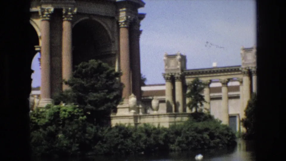 The Palace of Fine Arts appears from a visitor viewpoint across the water, with columns, arched architecture, trees, shrubs, and blue sky visible.