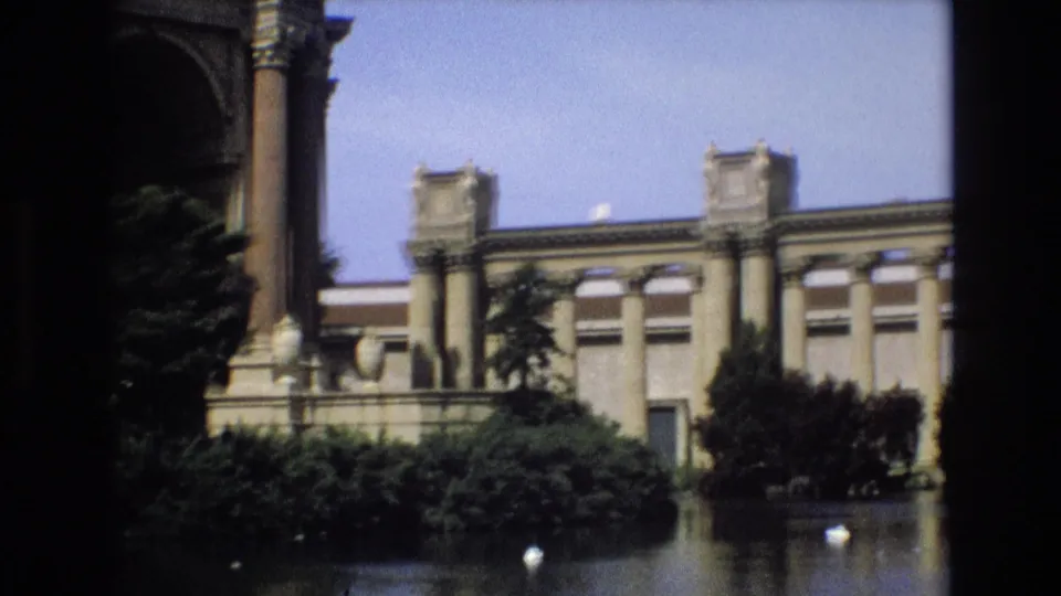 A visitor-shot 8mm frame shows the Palace of Fine Arts facade, trimmed plantings, reflecting water, and a small white bird near the lower center.