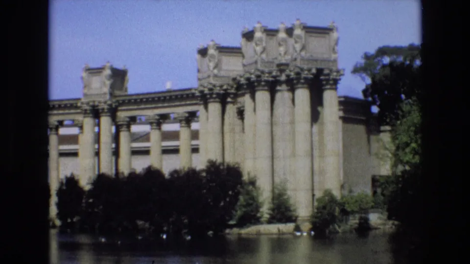 1973 San Francisco home-movie view of massive stone columns near a body of water, with dense shrubs and a leafy tree framing the architecture.