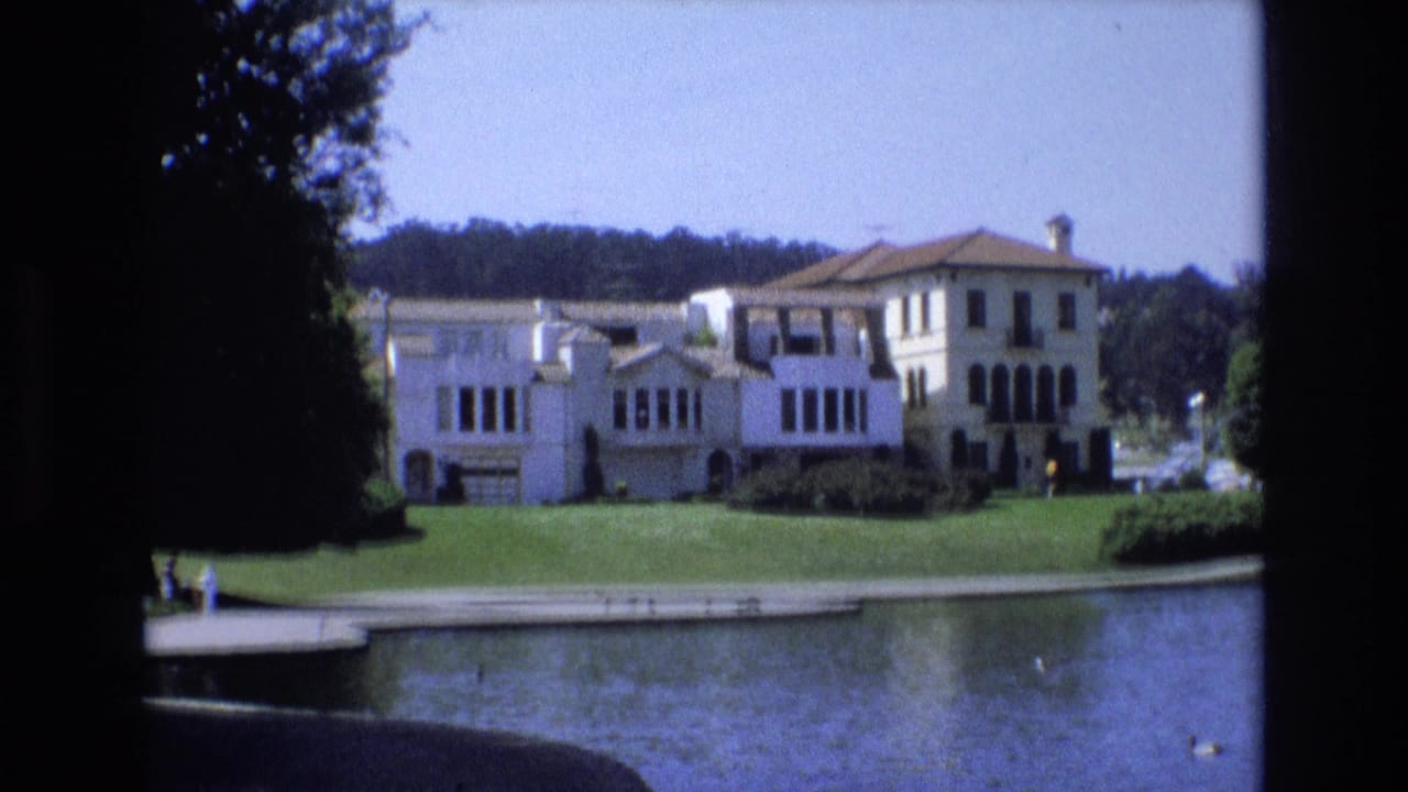 Pale villa-like building across a lake with lawn, trees, and small distant people.