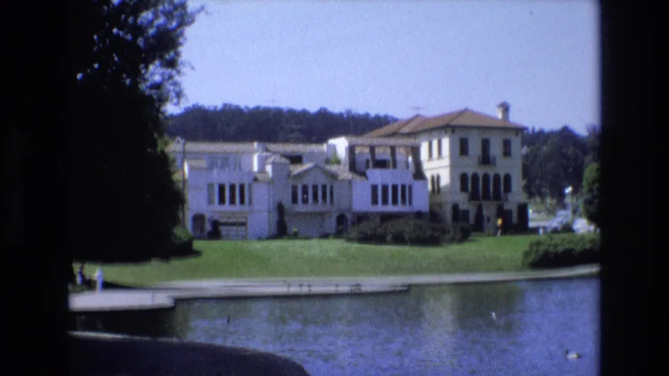 1973 San Francisco 8mm frame showing a pale Mediterranean-style building across a lake, with manicured grass, trees, and small distant visitors near the shore.