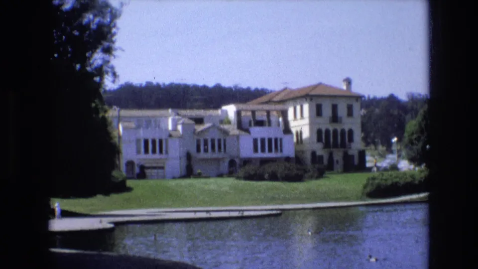 1973 San Francisco 8mm frame holding on a white villa-like building and green lawn across the lake, with dark trees framing the left edge and water in the foreground.