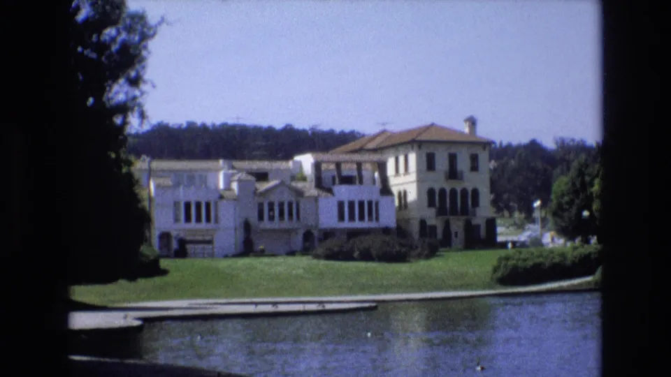1973 San Francisco 8mm frame showing the lakeside building, broad lawn, trees, and small ducks on the water in a steady home-movie view.
