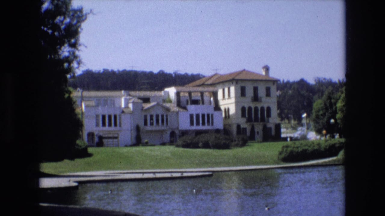 1973 San Francisco 8mm frame showing a white multi-story mansion across pond water, with a manicured lawn, dark windows, and trees around the property.