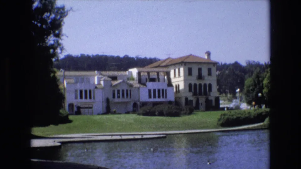 1973 San Francisco 8mm frame showing a white multi-story mansion across pond water, with a manicured lawn, dark windows, and trees around the property.